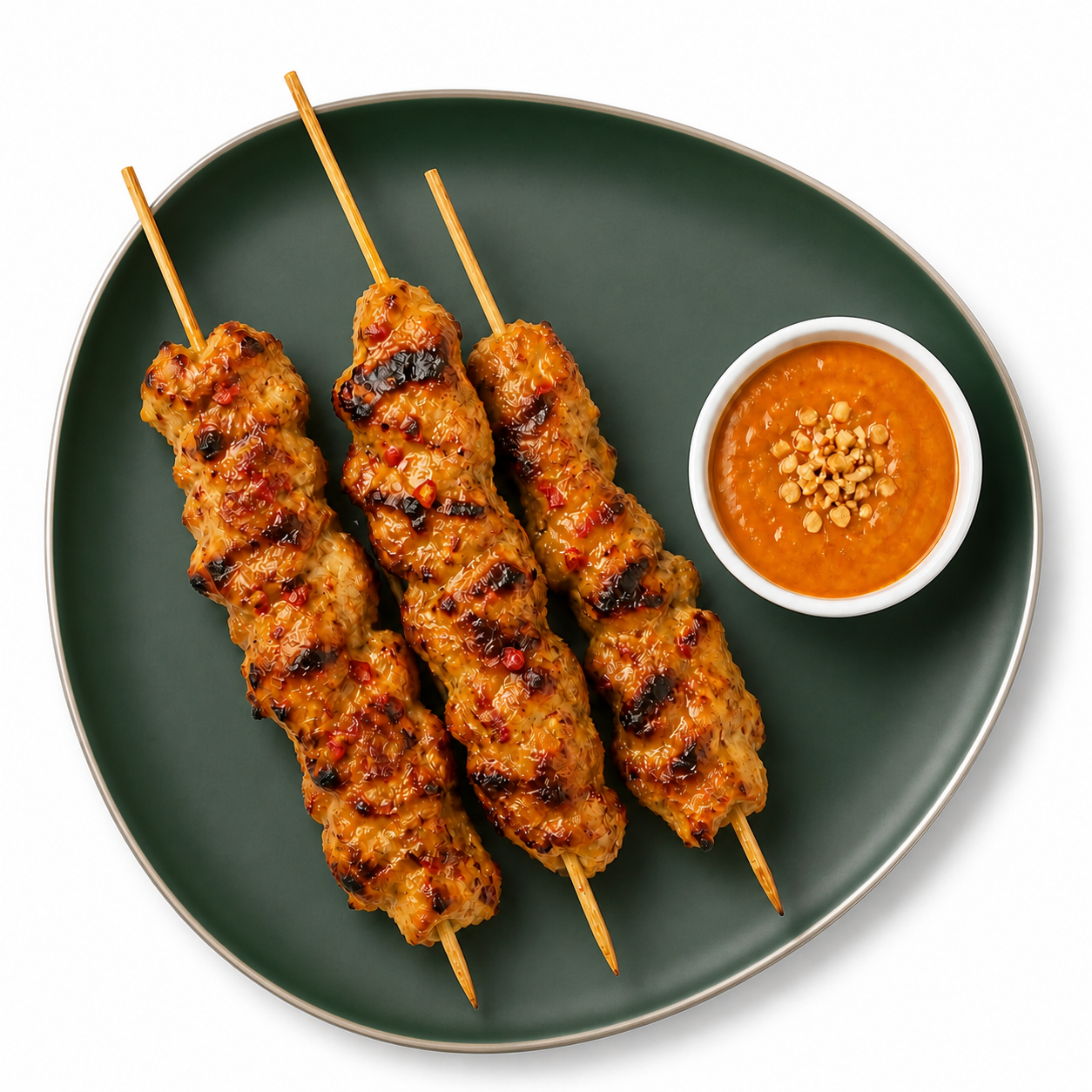 Three red-pepper-flecked chicken satay skewers with a one-ounce cup of red curry peanut sauce on a contemporary deep-green coupe plate